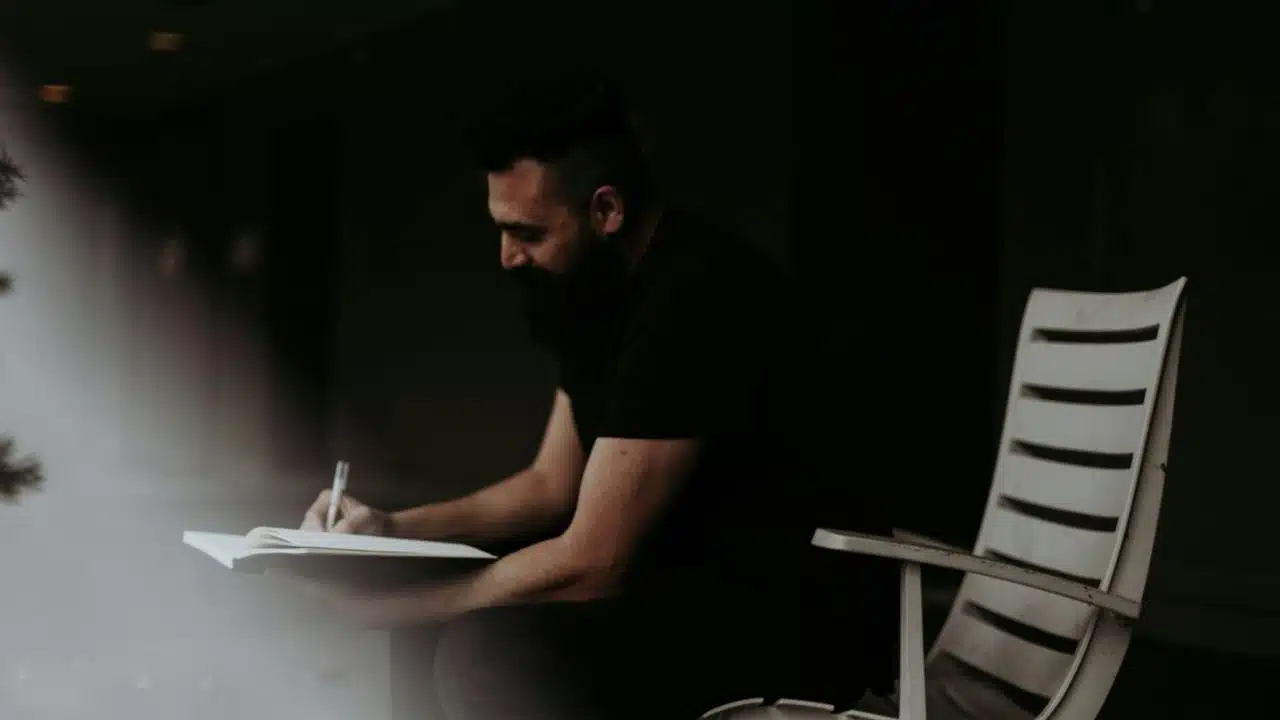 A focused man with a beard sits on a chair, writing in a book or journal.