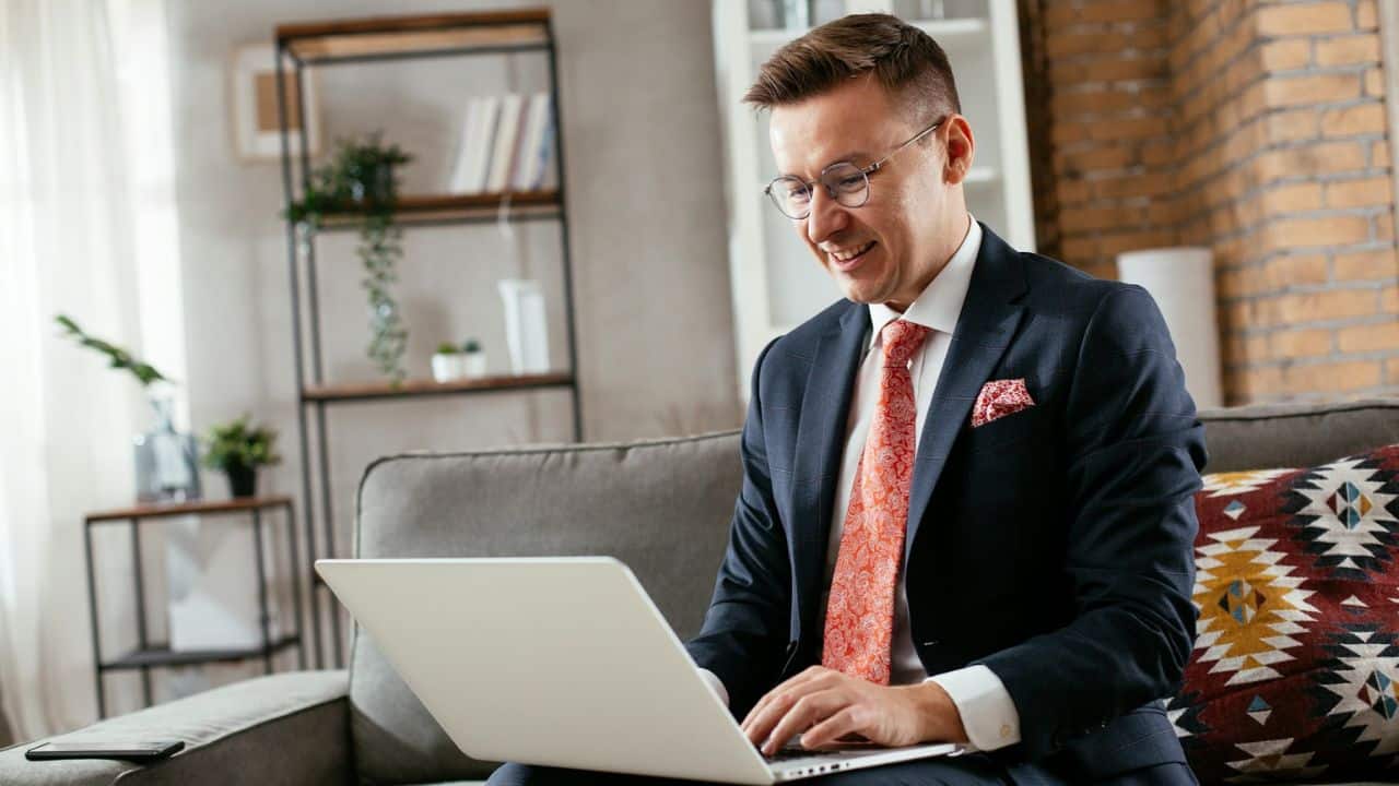 A professional-looking man in a suit jacket and tie is smiling and working on a laptop.