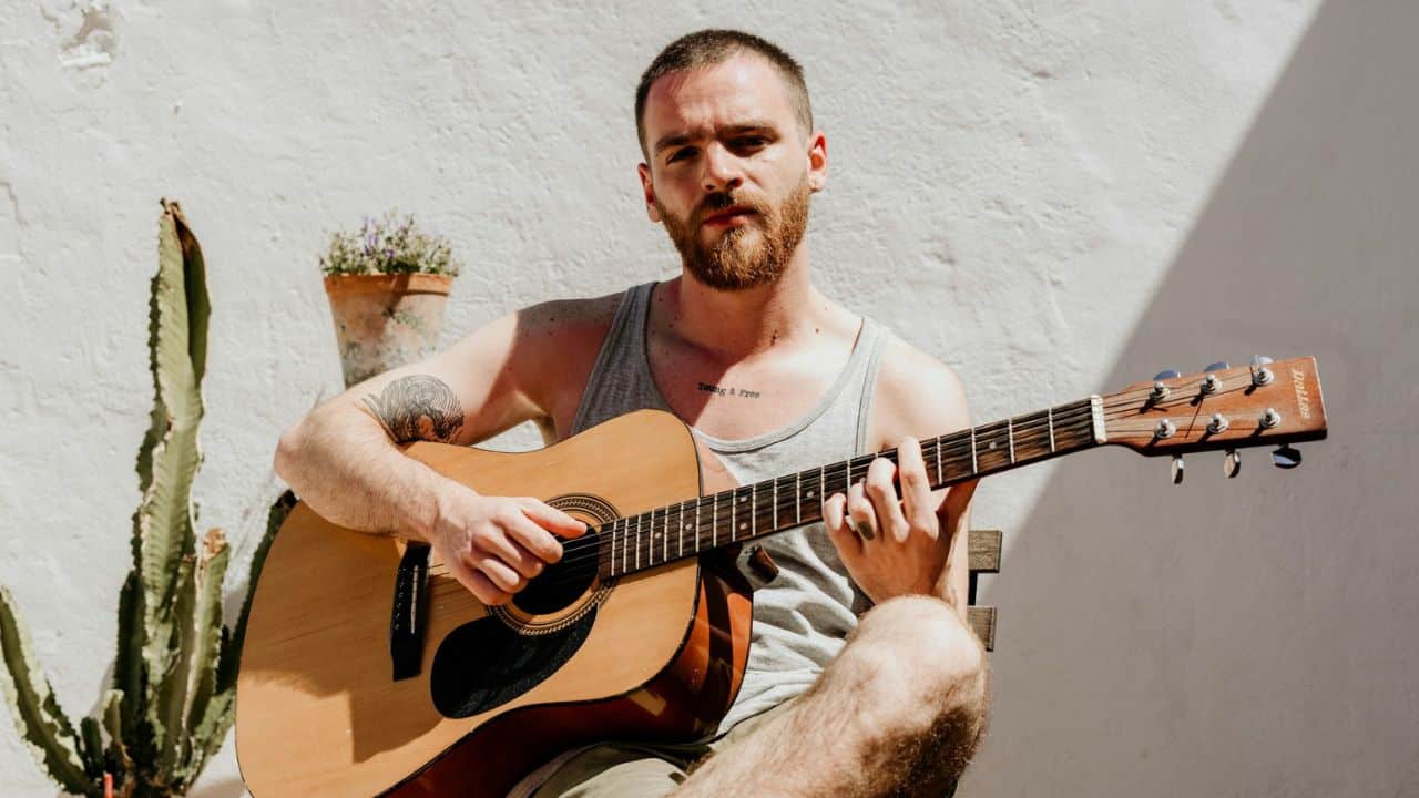 A bearded man in a tank top sits outside, holding and playing an acoustic guitar.