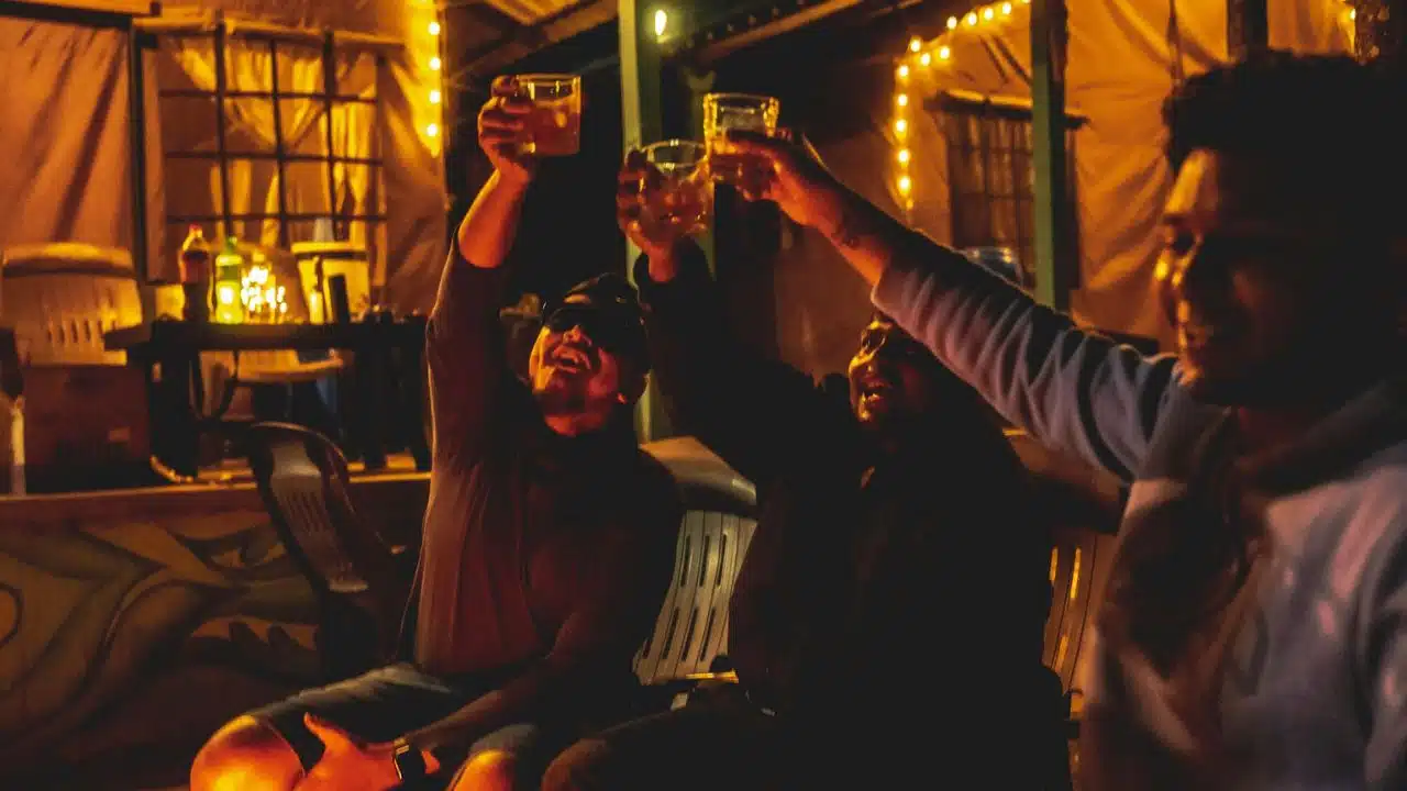 A group of happy friends sits together outdoors at night, toasting with drinks.