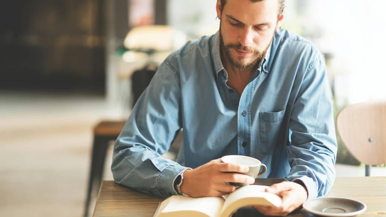 A bearded man in a denim shirt is sitting at a table with a cup, reading a book.
