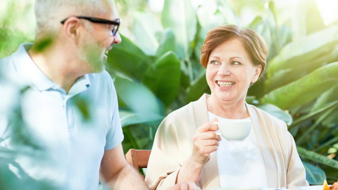 A happy, smiling couple sits together outdoors, with the woman holding a mug and looking at the man.