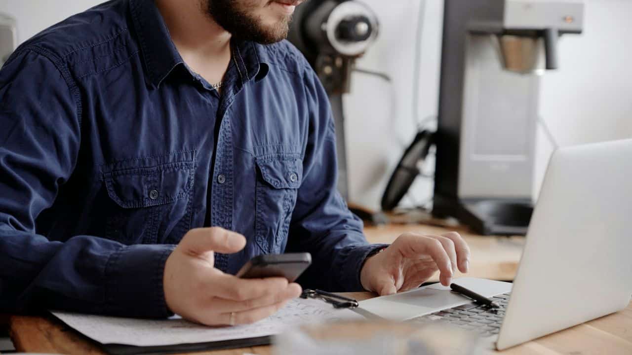 A man with a beard sits at a table, looking at his smartphone while also working on a laptop.