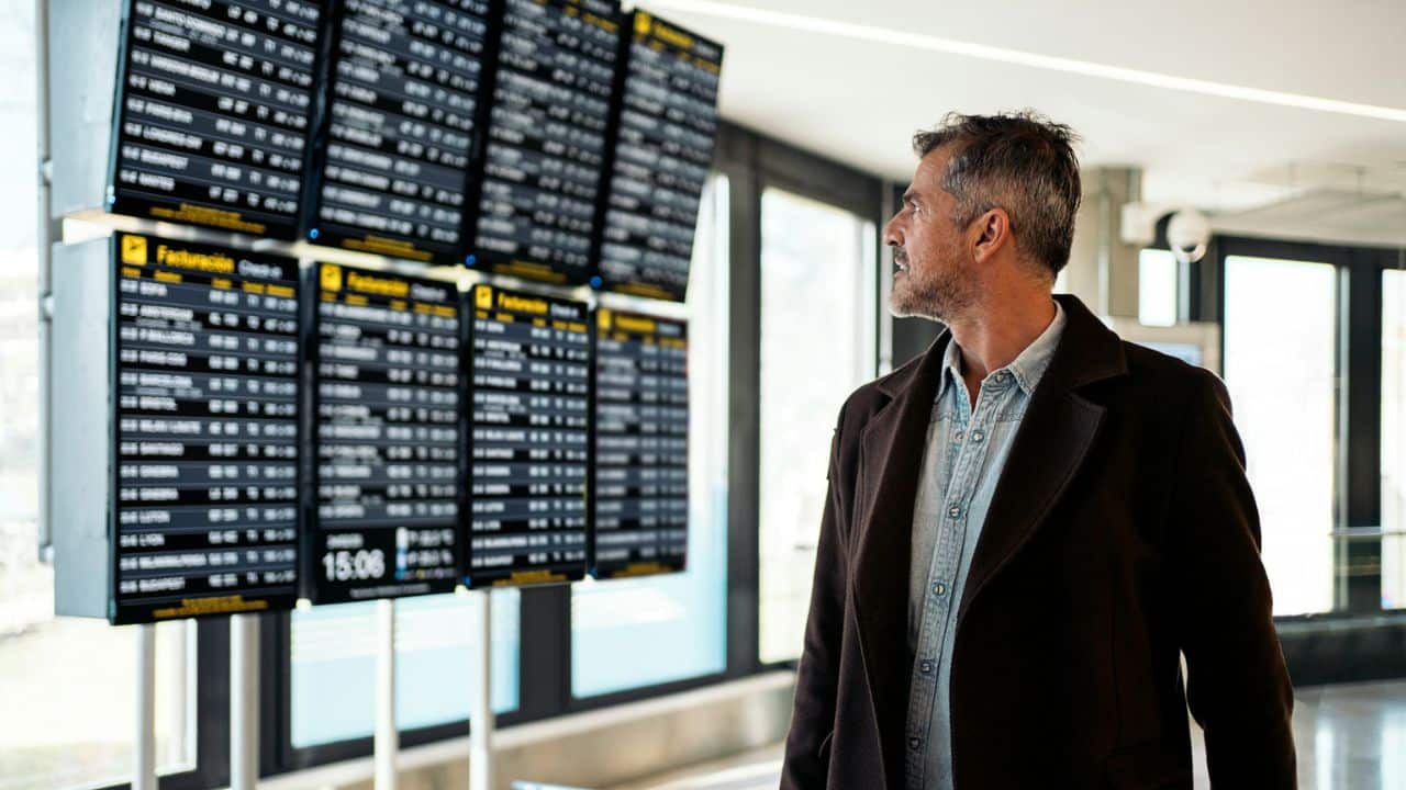 A happy man in a jacket looks at a departures board at an airport.