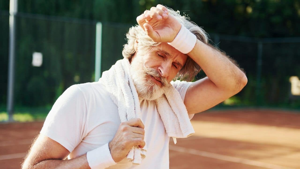 A sweaty, exhausted older man with a beard and a towel around his neck is standing on a tennis court, wiping his forehead.