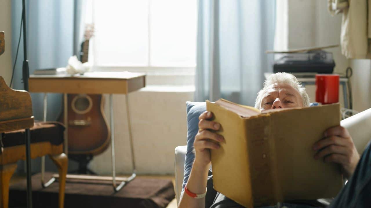 A happy, older man is lying on a couch in a sunlit room, reading a large book.