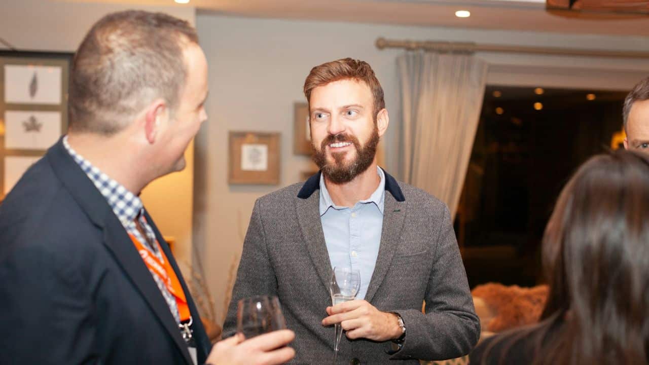 A handsome, bearded man is smiling and holding a champagne flute while talking to a friend at a gathering.