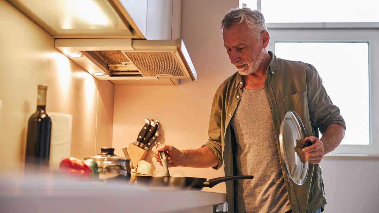 A handsome, older man with a beard is cooking in a kitchen, holding a pan lid and stirring something.