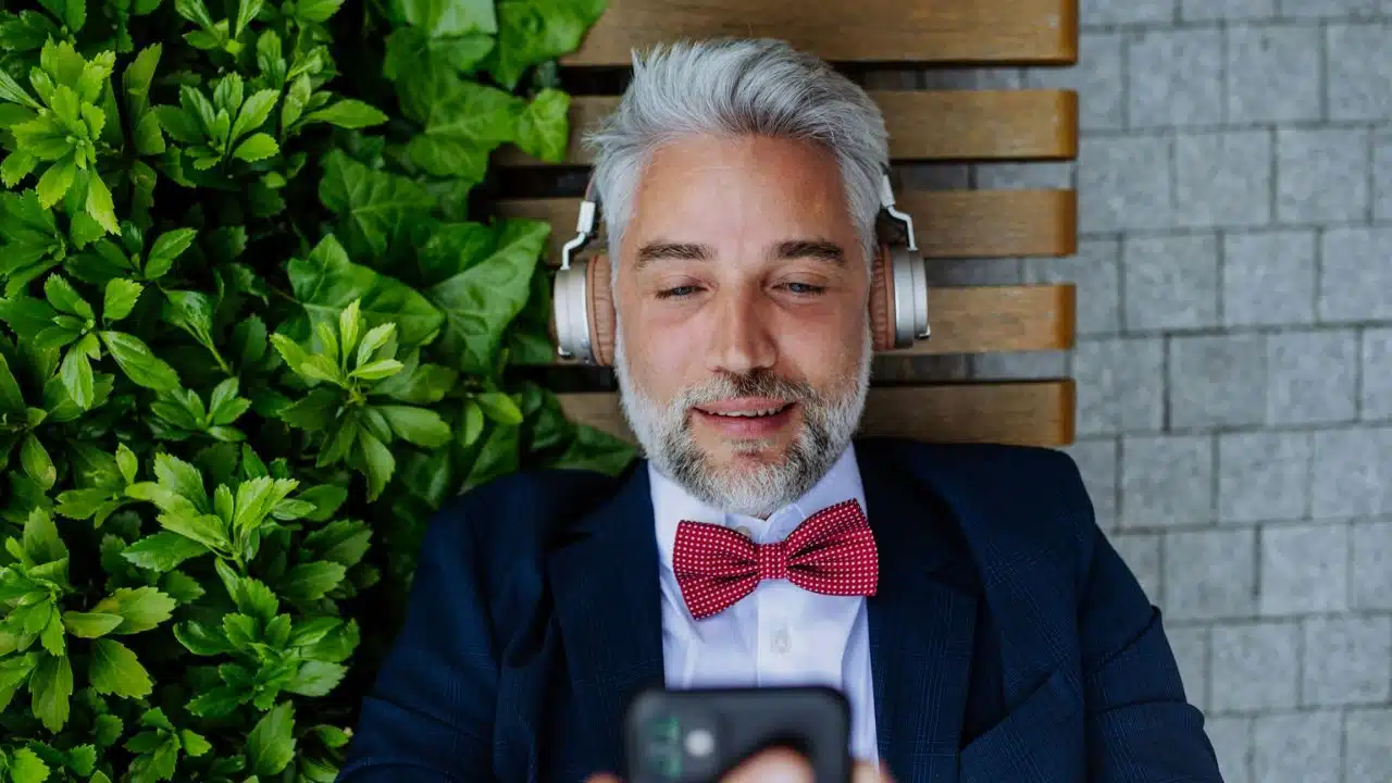 A handsome, older man with a beard and a bow tie is lying on a bench outdoors, wearing headphones and looking at his smartphone.
