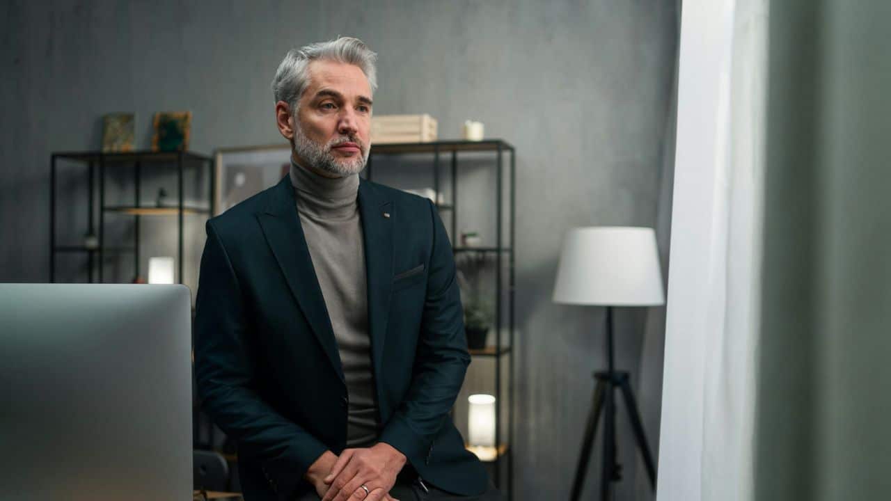 An elegantly dressed, middle-aged man with gray hair is sitting at a desk, looking off thoughtfully into the distance.
