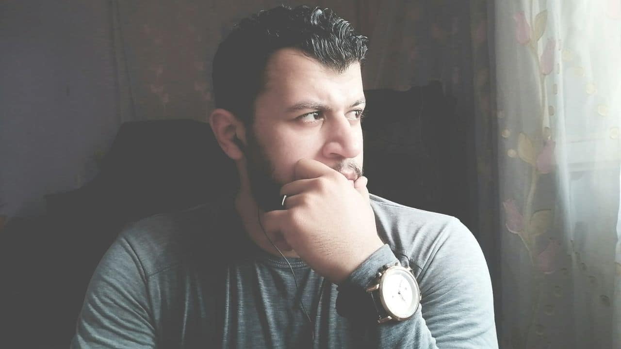 A bearded man in a gray shirt looks out a window, resting his chin on his hand.