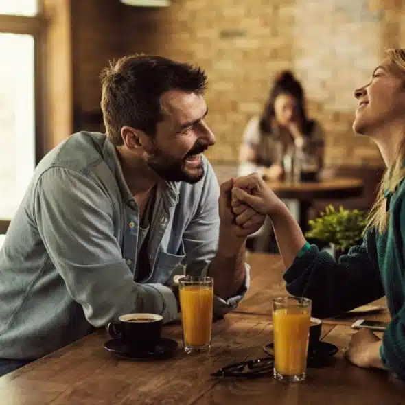 A happy couple is holding hands and laughing across a cafe table.