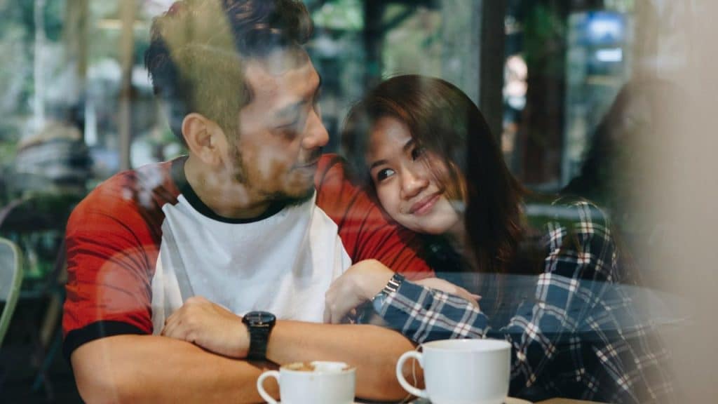 A smiling couple is leaning together at a cafe table with two cups of coffee.