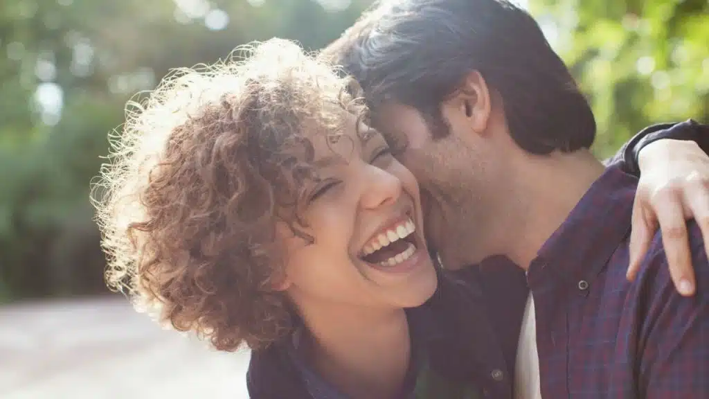 A laughing woman with curly hair is hugged by a man who is whispering in her ear.