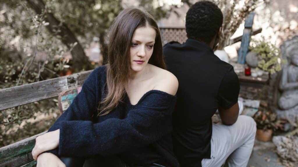 A solemn woman sits on a bench, turned away from a man who is also sitting on the bench.