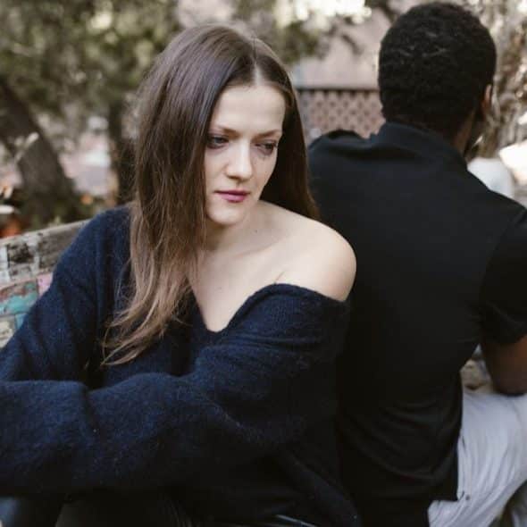 A solemn woman sits on a bench, turned away from a man who is also sitting on the bench.
