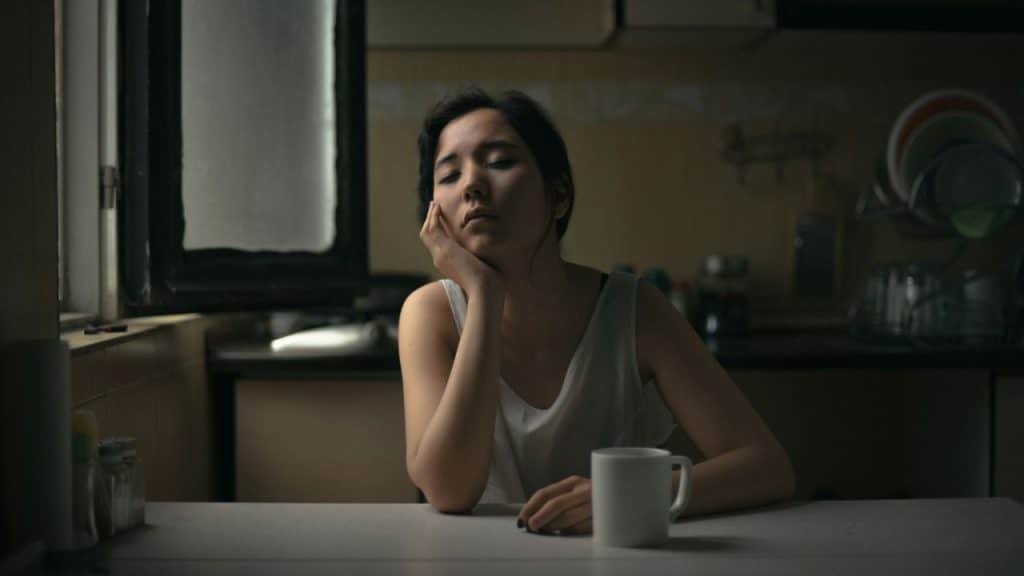 A weary-looking woman with her eyes closed sits at a kitchen table with a mug.