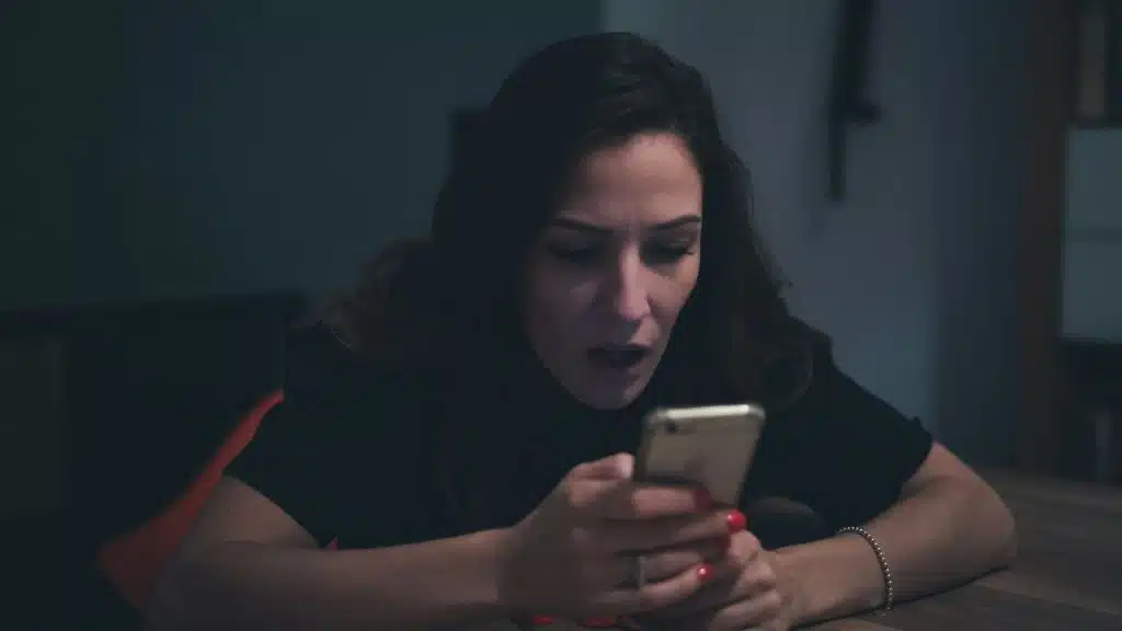 A shocked woman sits in the dark, looking at her smartphone with an open mouth.
