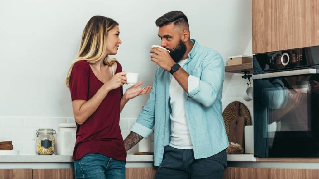 A woman is gesturing while talking to a man who is drinking from a mug in a kitchen.