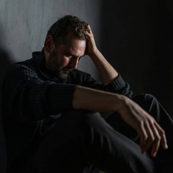 A distraught man sits in a dark room with his head in his hands.