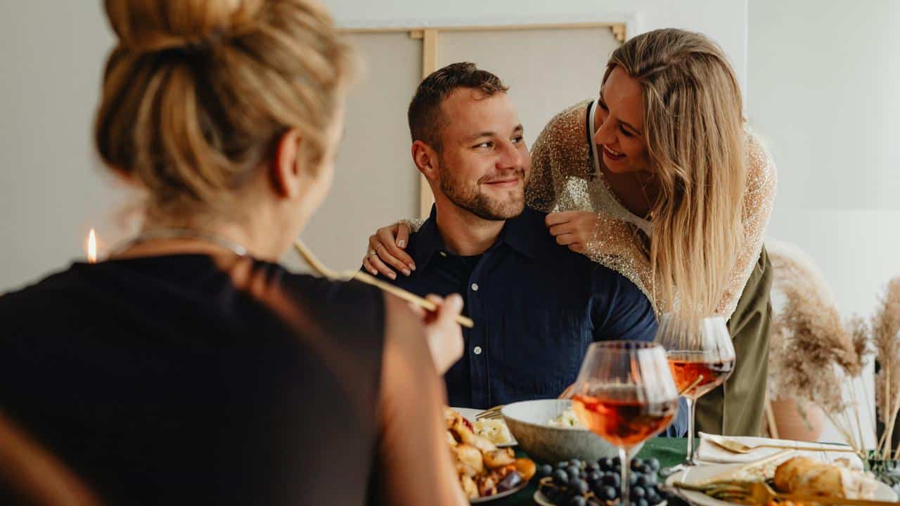 A woman leaning over a man’s shoulder as they smile during a meal with friends.