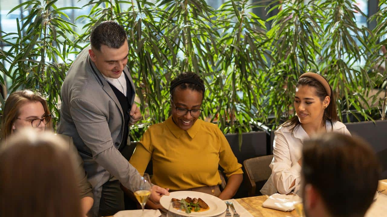 A waiter serving a smiling woman at a restaurant table with friends.