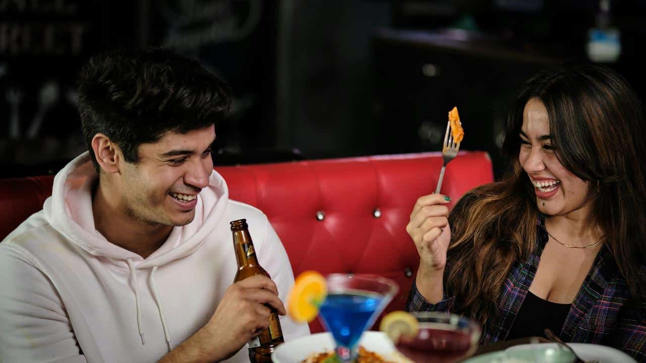 A man and woman laughing together while enjoying drinks and food at a restaurant.