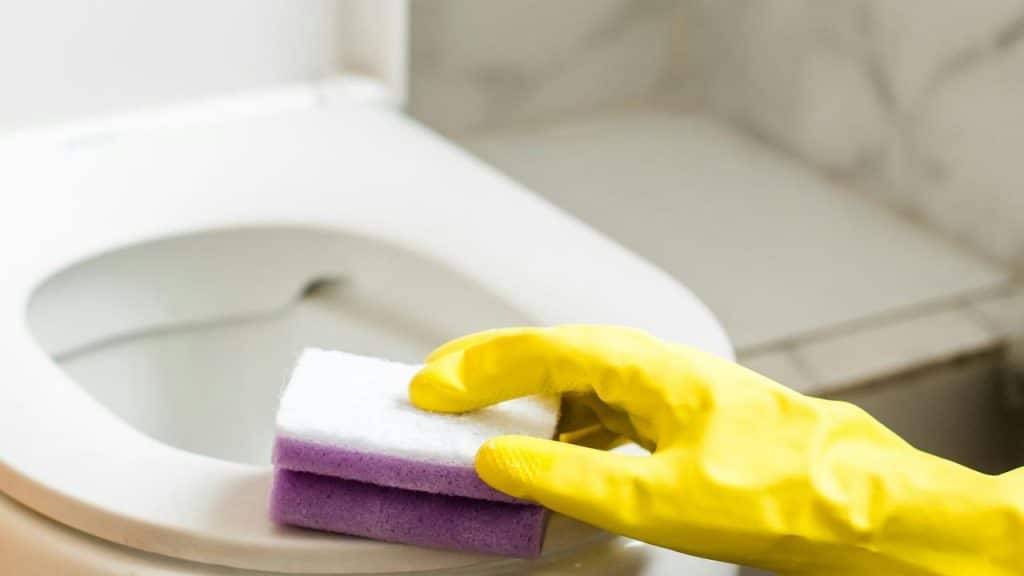 A person's hand in a yellow glove scrubbing a toilet with a sponge.