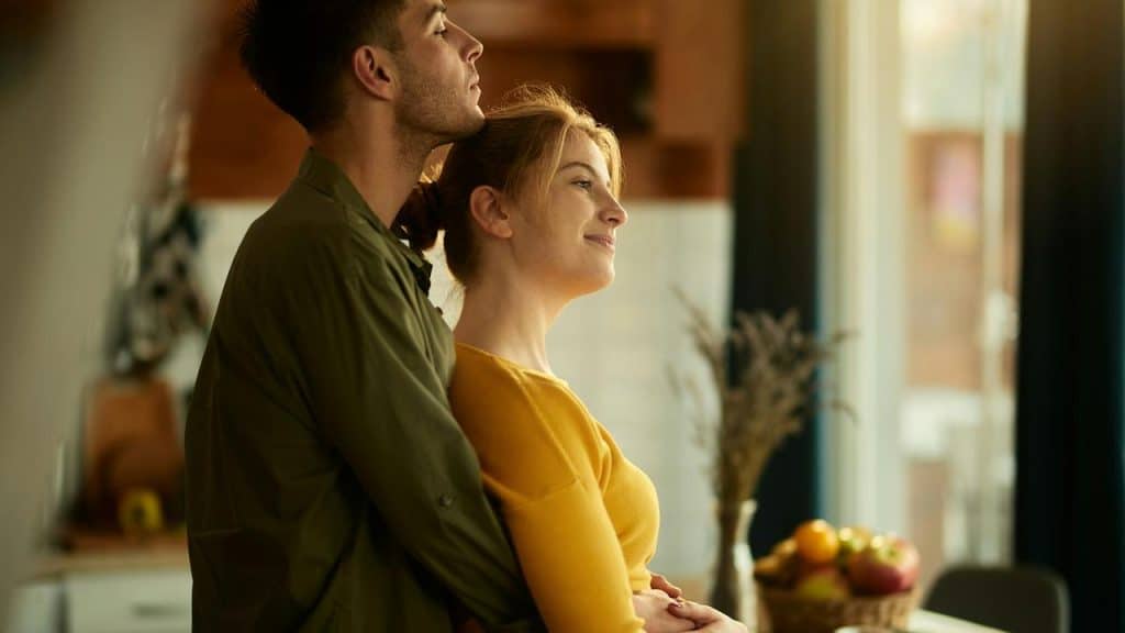 A man hugging a smiling woman from behind in a warmly lit kitchen.