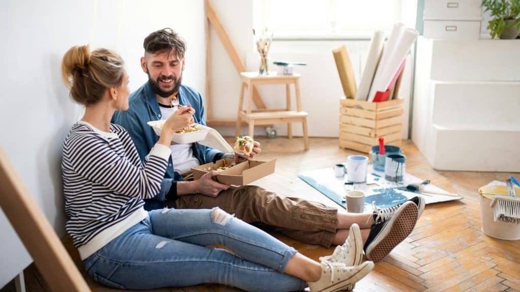 A couple sitting on the floor eating takeout amid home renovation supplies.