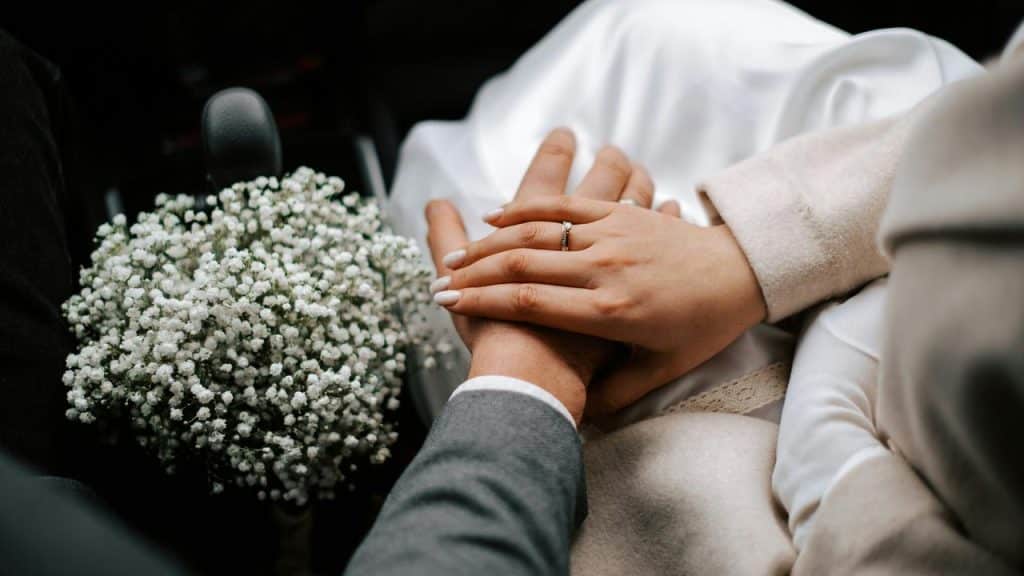 A couple holding hands with a wedding ring visible and a bouquet of white flowers nearby.