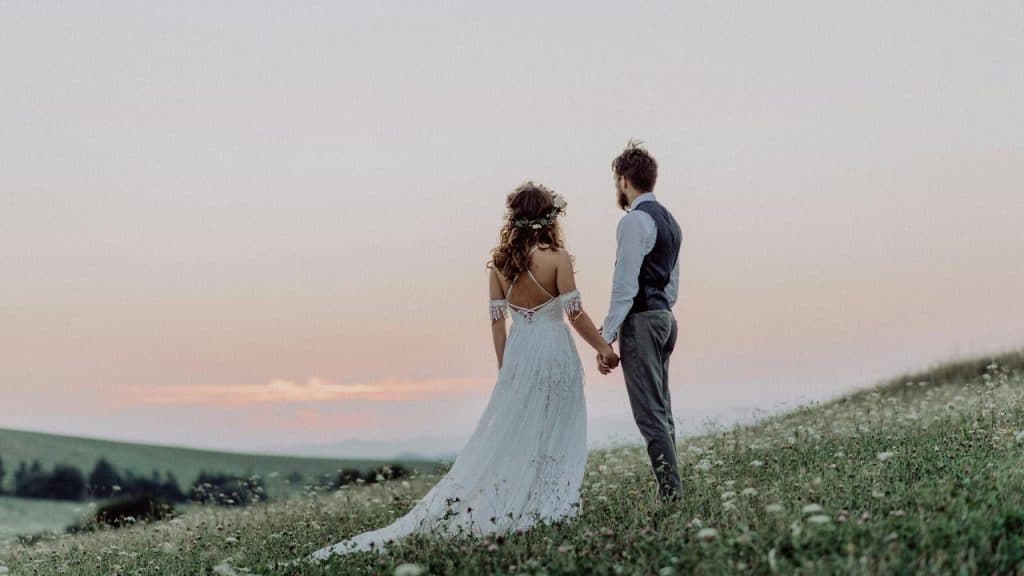 A bride and groom holding hands while standing in a field at sunset.