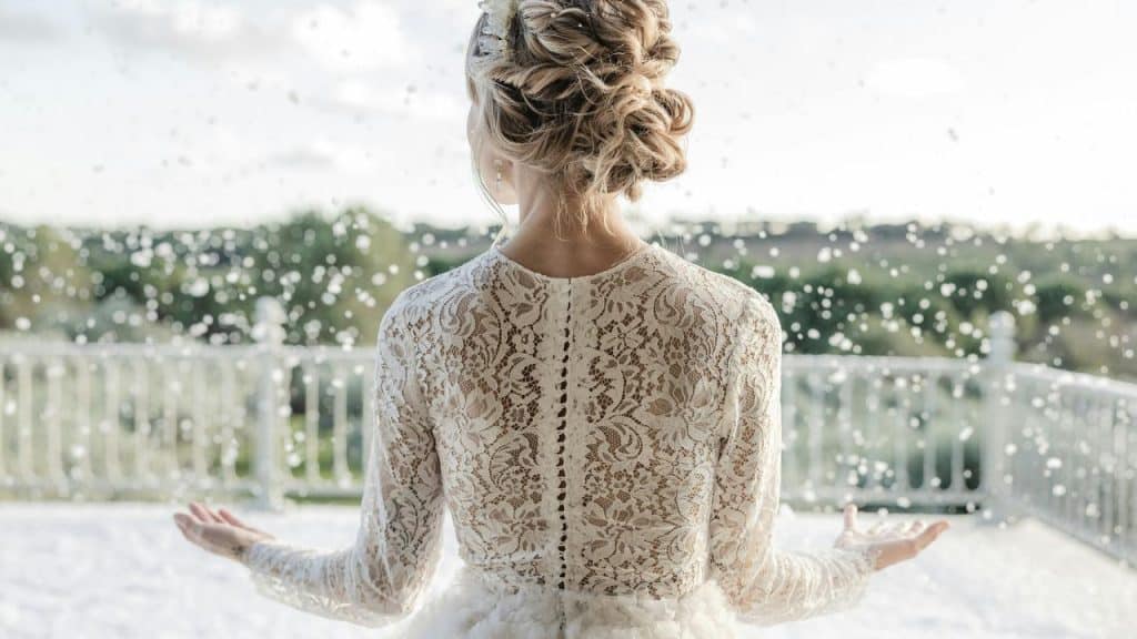 A bride in a lace dress standing outdoors with arms open as petals fall around her.