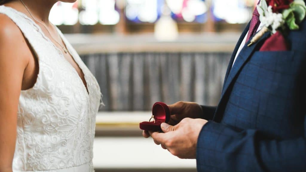 A groom holding an open ring box in front of a bride during a wedding ceremony.