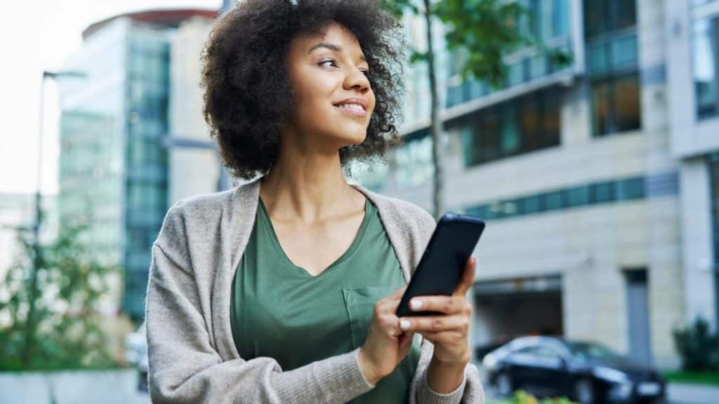 A woman holding a smartphone and smiling while looking away outdoors.