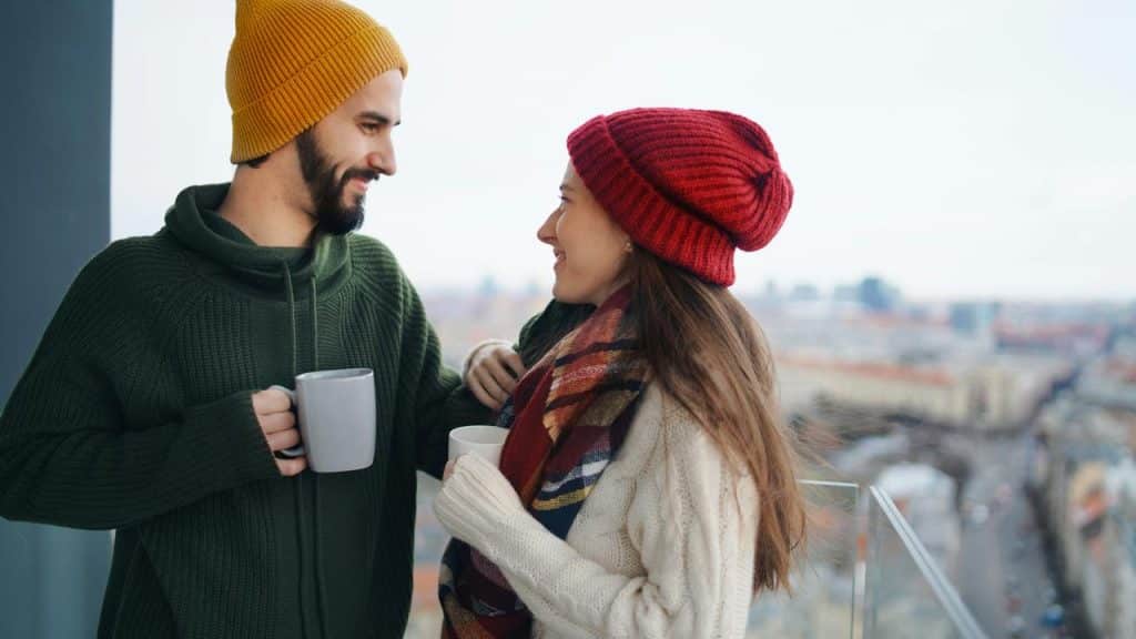 A couple in winter clothing smiling at each other while holding mugs on a balcony.