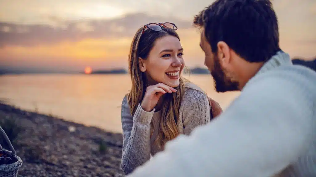 A couple smiling and talking by the water during sunset.