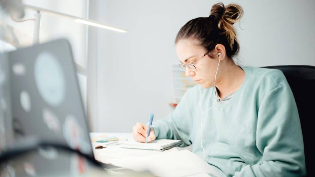 A woman writing in a notebook while using a laptop.