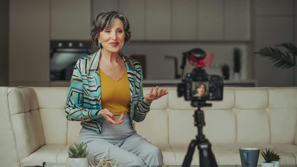 An older woman speaking on camera while sitting on a couch.