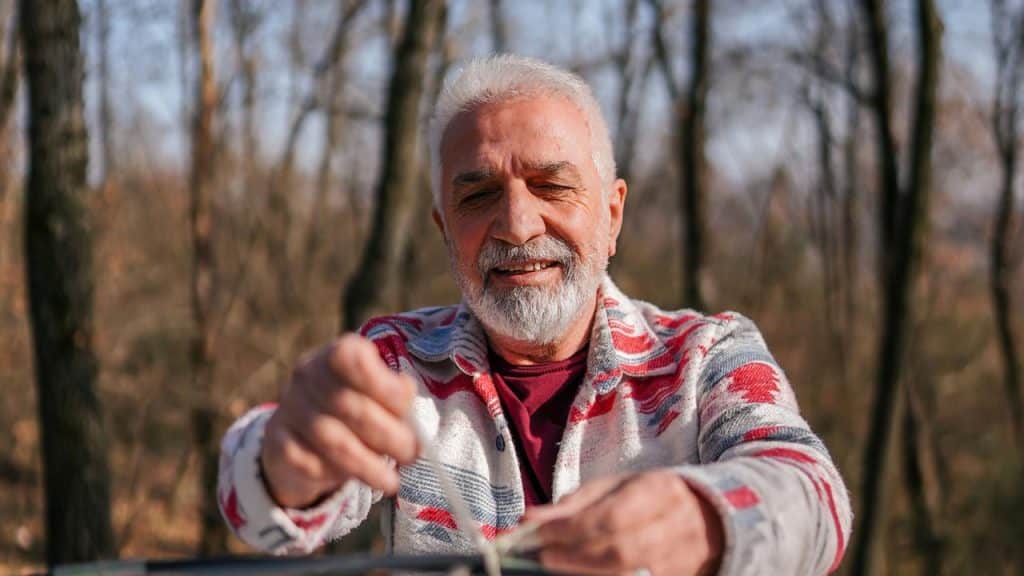 An older man smiling while tying a rope outdoors in a forest.