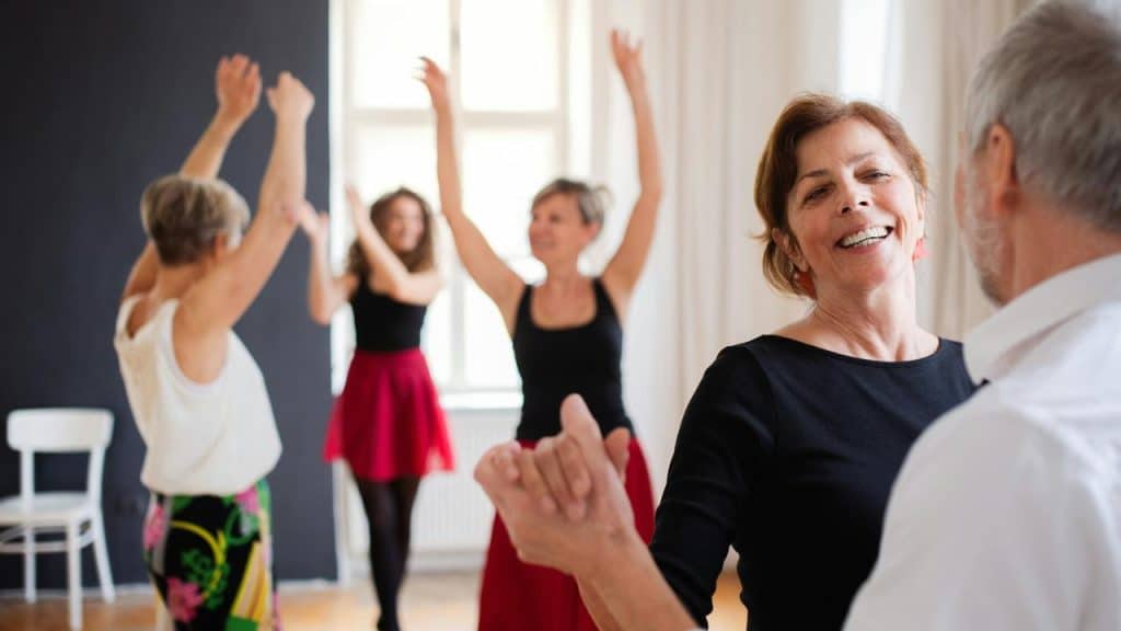 An older woman smiling while dancing with a partner in a group dance class.