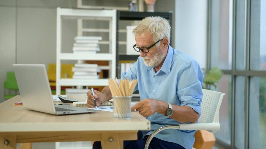 An older man with glasses working at a desk with a laptop and papers.