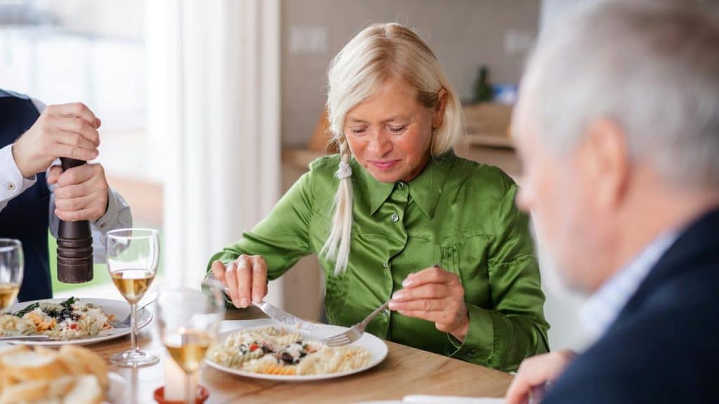 An older woman in a green shirt eating pasta at a table with others.