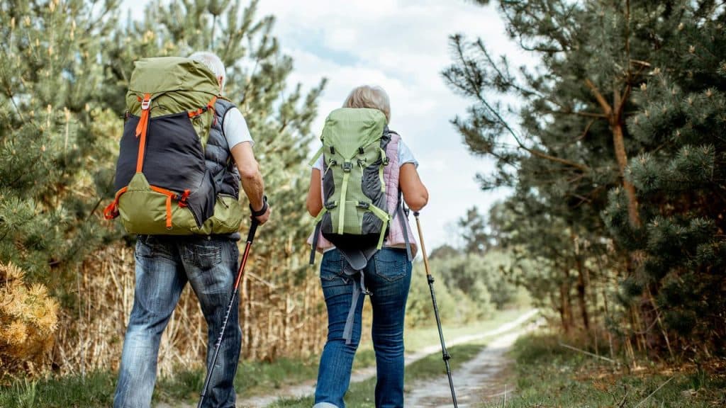 An older couple hiking on a forest trail with backpacks and walking sticks.