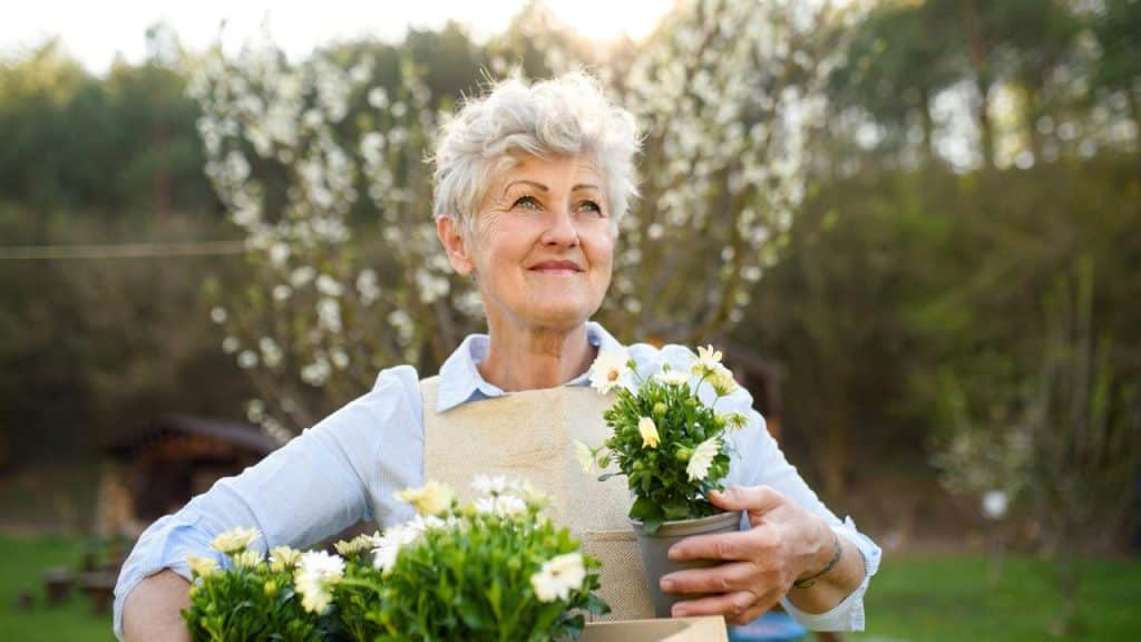 An older woman holding potted flowers in a garden.
