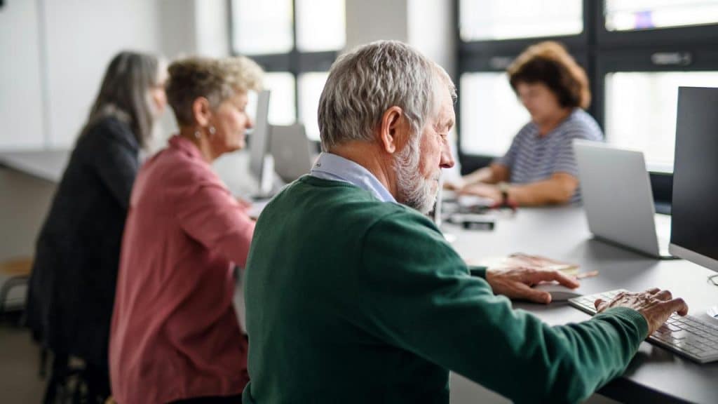 An older man using a computer in a classroom with others.