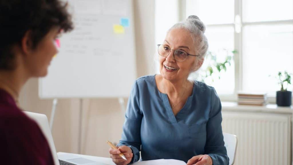 An older woman with glasses smiling while talking to someone in an office.