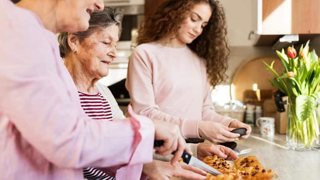 An elderly woman and two others preparing food together in a kitchen.