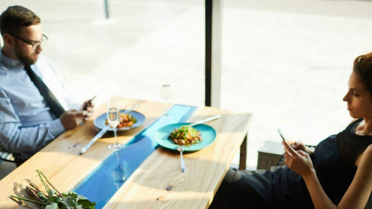 A man and woman sit at a table, both looking at their phones.