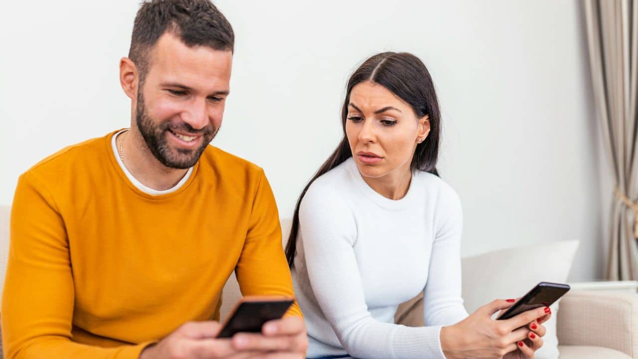 A smiling man looks at his phone while a woman next to him looks worried.